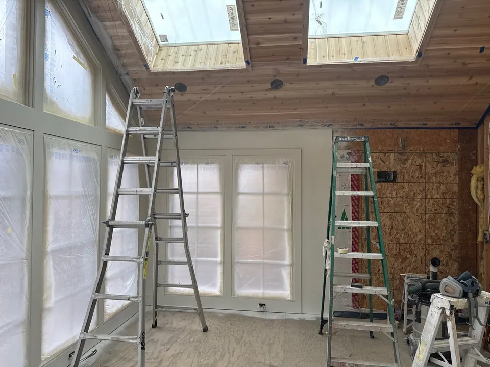 Room mid-paint with the windows masked in plastic, two ladders set up under a wood ceiling with skylights, and the walls primed white.